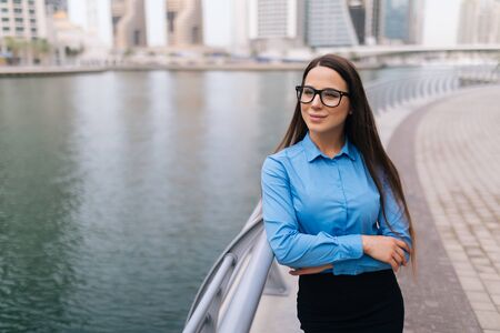 Portrait of young beautiful business woman in suit, glasses, smiling, successful looking at sides, with skyscraper background.の写真素材