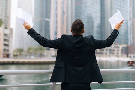 Portrait of handsome young businessman with raised hands holding sign contract celebrate against view of skyscrapersの写真素材