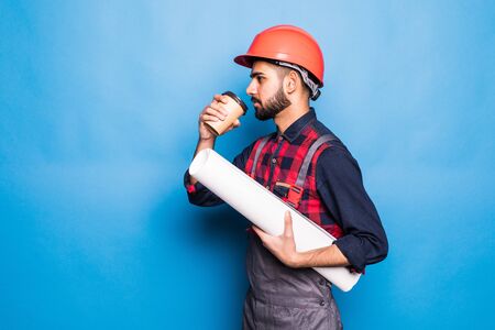 Smiling indian engineer in safety vest holding blueprints and coffee to go on blue backgroundの写真素材