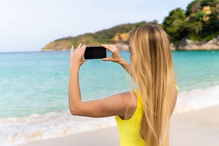 Back view of a woman taking photograph with a smart phone camera on the beachの写真素材