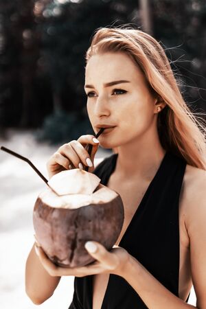 Tropical vacation. Young woman in hat and swimsuit holding green cococnut relaxing under coconut palm.の写真素材