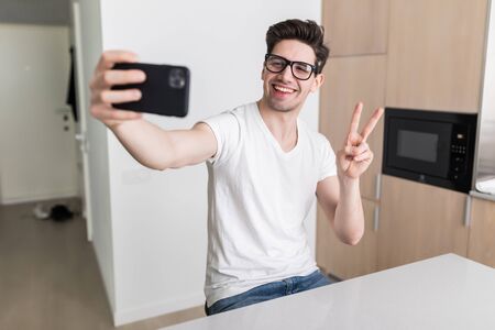 Young man taking selfie and making peace gesture at homeの写真素材