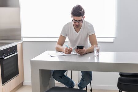 Young man is writing something on paper notebook and holding mobile smart phone at home. business concept.の写真素材