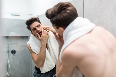Young man standing in front of mirror and touching his faceの写真素材