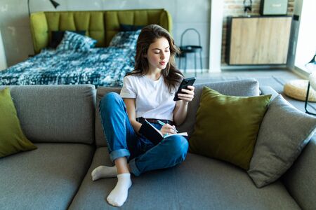 Close up of smiling woman sitting near sofa, writing in weekly planner and surfing phone. Working at home.の写真素材