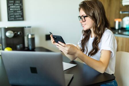 Young woman with laptop using credit card and phone in kitchen at home