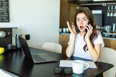 Beautiful shocked girl is talking on the mobile phone, using a laptop at home