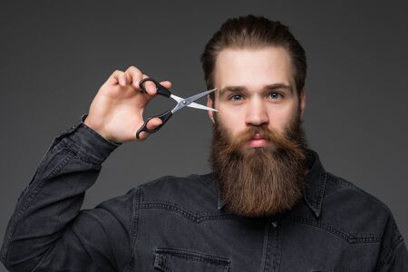 Young bearded man holding Barber scissors and razor near his face standing on gray background
