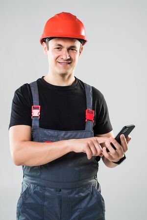 Portrait of young male construction worker with phone wearing protective clothes, helmet and tool belt isolated on white backgroundの写真素材