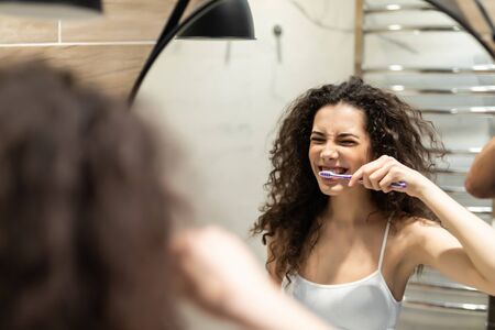 Portrait of attractive woman brushing teeth in bathroom and looking in the mirror at reflection.