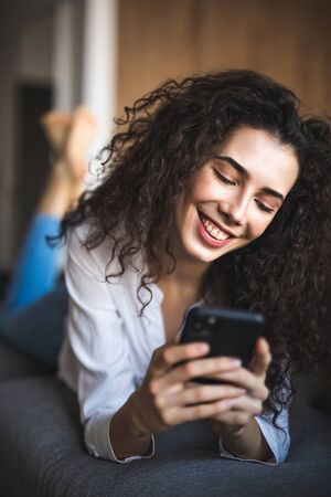 Happy woman lying on the couch at home, talking on the phone with friend
