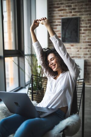 Close up of young cute smiling woman sitting on modern chair near the window in light cozy room at home, working on laptop with win gestureの写真素材