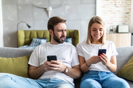 Young couple using smartphones at home. Jealous boyfriend spying his girlfriend's phone.の写真素材