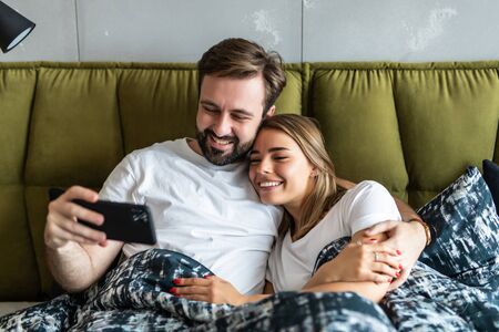Smiling attractive millennial couple using smartphone lying on bed togetherの写真素材