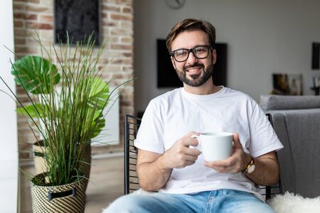 Young man drinking a cup of coffee while sitting on a chair at homeの写真素材