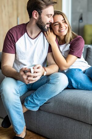 Beautiful young couple is talking and smiling while sitting on sofa at home.