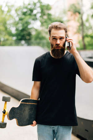 Young man with a skateboard is talking by a mobile phone while sitting in a skate park on a sunny day.の写真素材
