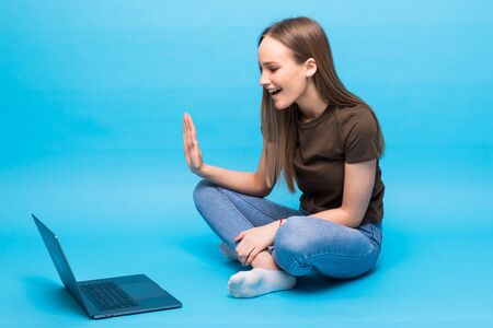 Young woman waving by hand and having video call on laptop sitting on the floor on blue background