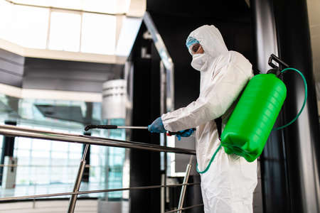 Professional fully armed disinfector against Covid-19 using sprays to remove bacteria from the surface at the hotel. Man wearing a protective mask, gloves and suit.の写真素材
