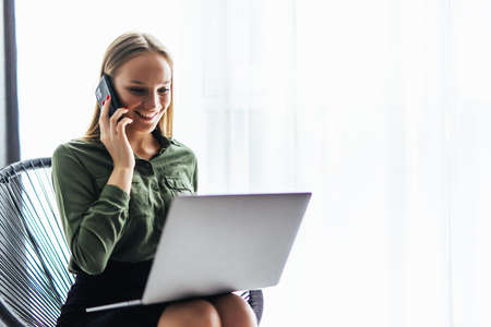 Enjoying time at home. Beautiful young smiling woman working on laptop and make a call while sitting...