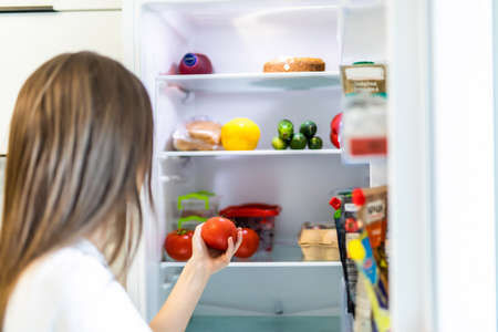 Young pretty woman taking fresh healthy tomato vegetables from refrigerator, view from fridgeの写真素材