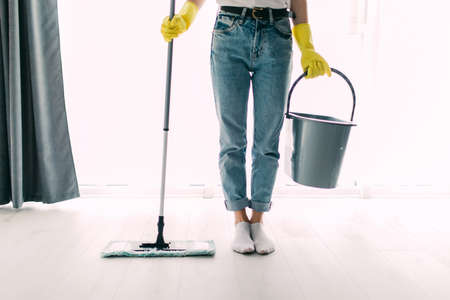 Happy young woman in blue rubber gloves wiping dust using mop while cleaning on floor at home.