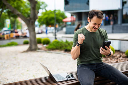 Happy man celebrate win look at phone sitting on a bench in the parkの写真素材