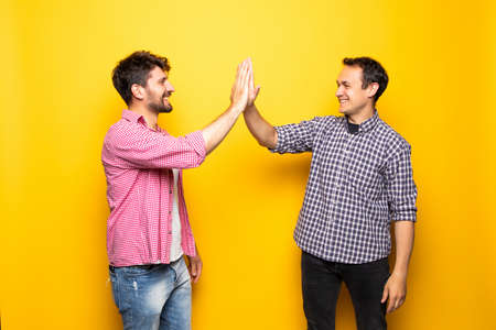 Portrait of a two delighted young men celebrating with high five gesture isolated over yellow backgroundの写真素材