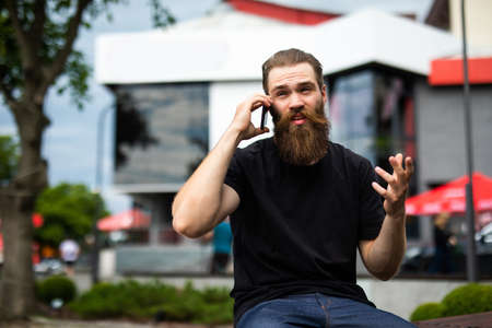 Handsome smiling young bearded man wearing jacket sitting on a bench and talking on mobile phone while looking at documentsの写真素材