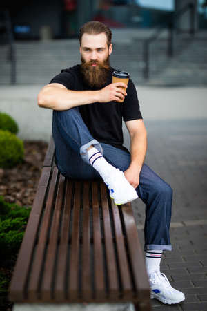 Image of handsome adult man in jacket smiling and drinking coffee while sitting on bench in parkの写真素材