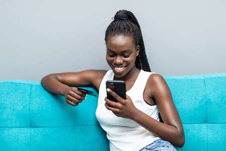 African woman at home couch using credit card for buying and banking online with mobile phone sitting on couch at homeの写真素材