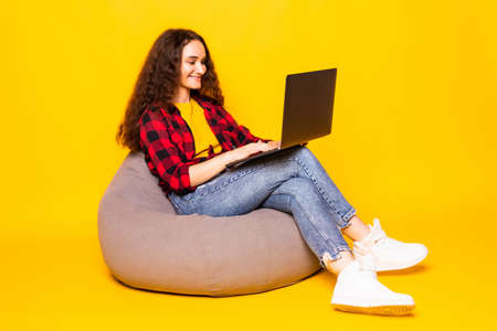 Young smiling woman sitting on floor with laptop on yellow backgroundの写真素材