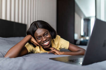 Beautiful african american professional woman laying relaxing on hotel bed, using a laptop computer, reading serene, travel interior.の写真素材