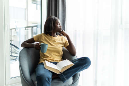 people and leisure concept - happy smiling african american woman sitting on sofa and reading book at homeの写真素材