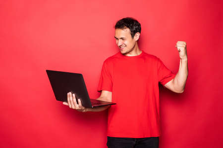 Portrait of a cheerful young man in red t-shirt holding laptop computer and celebrating isolated over red backgroundの写真素材
