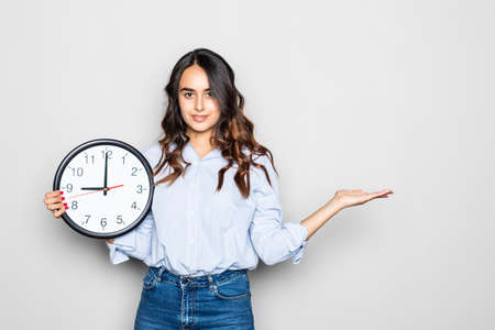 Smiling beautiful young girl holding in hand clock in white shirt posing isolated on gray wall...