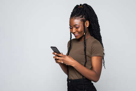 Happy young african woman casually dressed standing isolated over gray background, holding mobile phoneの写真素材