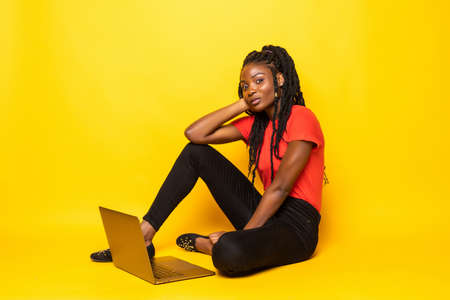 Online order. Happy afro woman using laptop, talking on phone while sitting on floor over yellow...