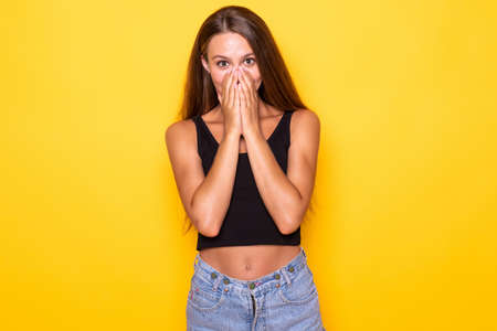 Young woman covered her mouth with both hands, shocked on a yellow background.