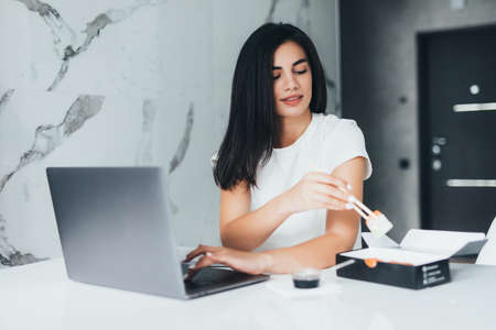 Young woman sitting at her kitchen eat sushi at table home working with laptopの写真素材