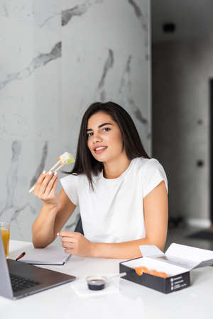 Young woman sitting at her kitchen eat sushi at table home working with laptopの写真素材
