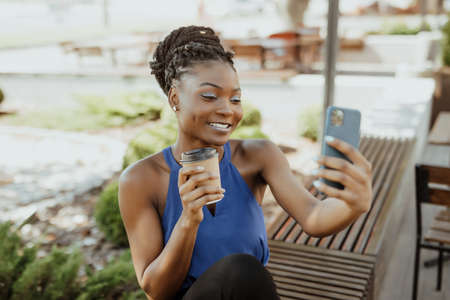 Cheerful african woman walking outdoors, holding takeaway coffee cup, taking a selfie sitting on the...