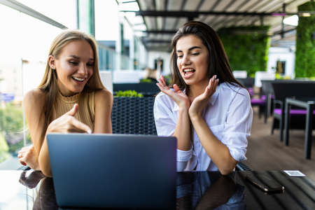 Two young women wearing in shirts are sitting in cafe at table and using laptop, working, blogging, online shopping.の写真素材