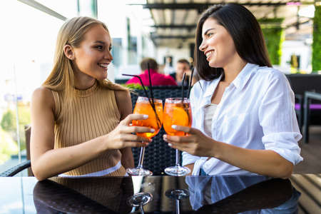 Young women drinking aperol spritz, clink glasses with friends, looking at camera in cafeの写真素材