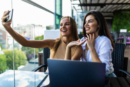 Two young attractive women making selfie photos on telephone sitting in coworking space with laptop computer.Charming students dressed in casual outfit making pictures on front camera of cellularの写真素材