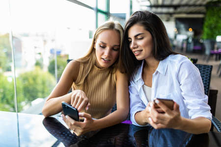 Two cute women in casual clothes are looking at the screen of a mobile phone while sitting in a street cafe.の写真素材
