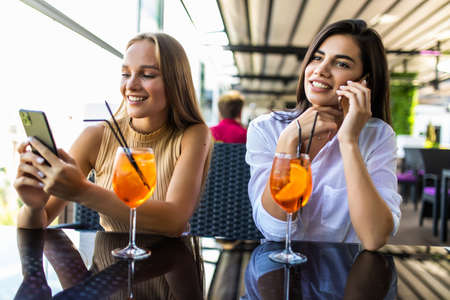 Positive young women recreat together at cozy restaurant, use phones, enjoy tasty cocktails. Multiracial relationship conceptの写真素材