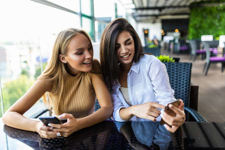 Two cute women in casual clothes are looking at the screen of a mobile phone while sitting in a street cafe.の写真素材