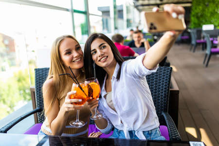 Two young woman take a selfie while sitting and chilling in the summer terrace of city cafeの写真素材