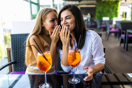 Two women sharing secrets and covering faces while drink cocktails in cafe.の写真素材